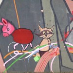 Das Foto zeigt ein Teil eines Wandgemäldes, auf dem ein roter Esel mit rosa Kopf uind ein kleiner Waschbär zu sehen sind