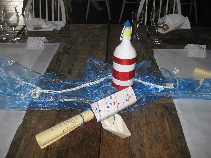 Eine als Leuchtturm bemalte Flasche steht auf blauem Organza, daneben steht ein Schiffchen, das die Menükarte hinter sich herzieht. 