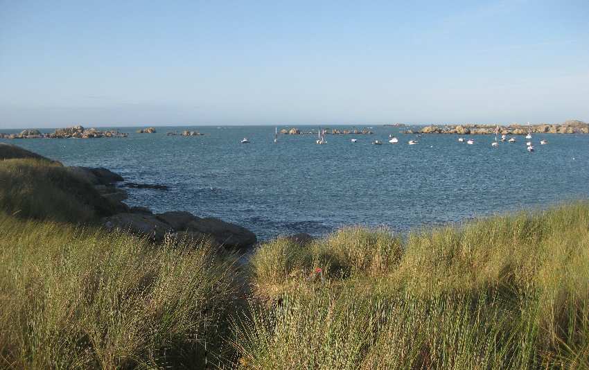 Blick auf die Bucht, im Vordergrund das Gras der Dünen, im Hintergrund einige große Felsen, dazwischen ein kleiner Naturhafen mit einigen kleinen Booten
