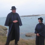 Joel und Marie-Pierre auf den Klippen vor Kerlouan, beide tragen dunkelblaue Mäntel und schwarze Hüte, die Kleidung der Conteurs