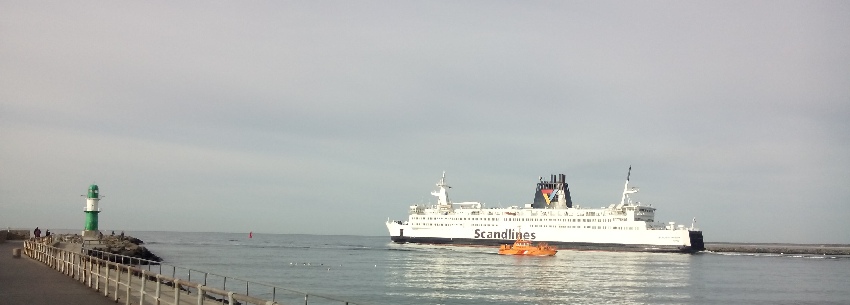 Große Fähre der Scandlines fährt in den Hafen ein, davor ein orangfarbenes Schlepperboot